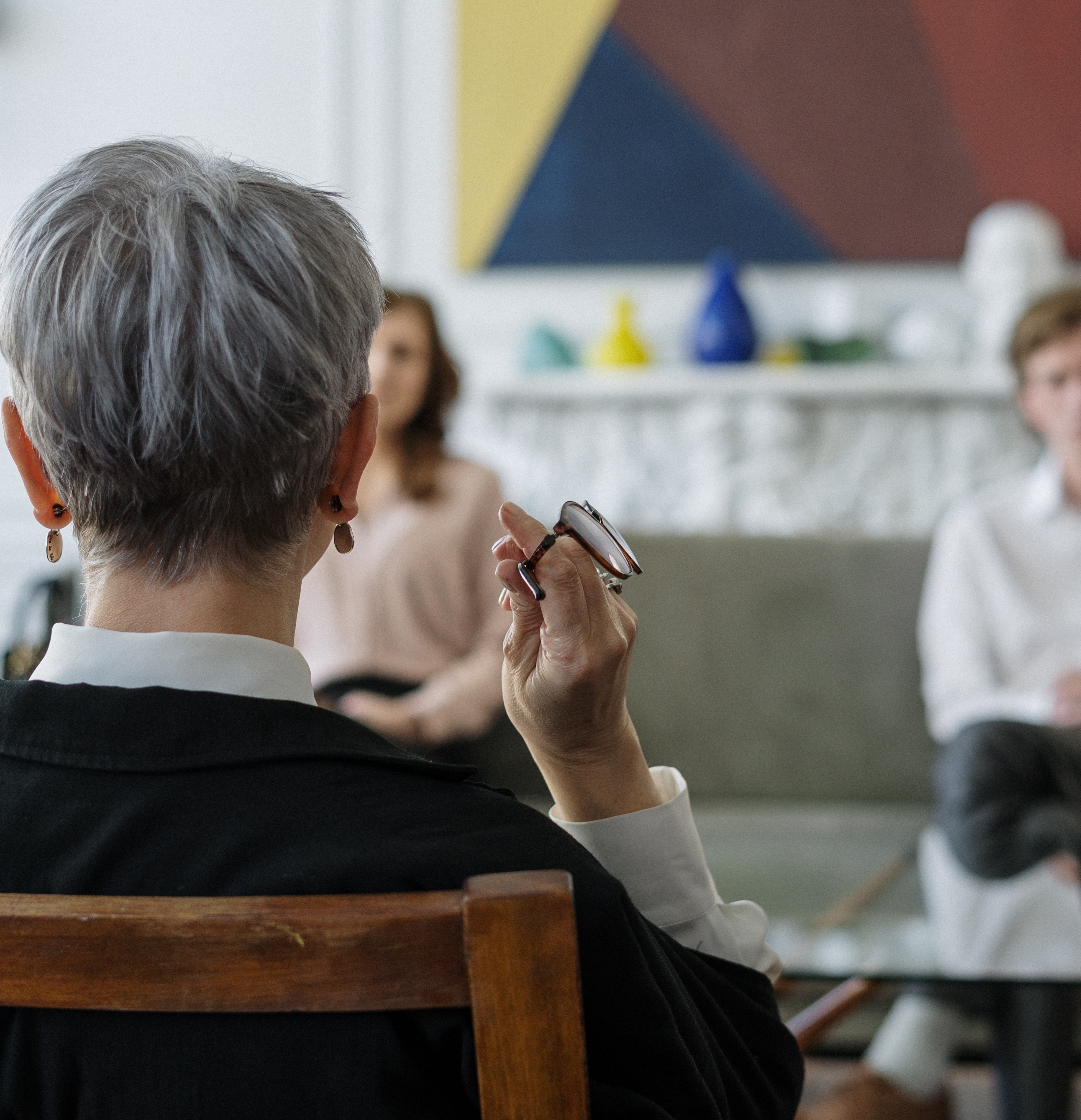 Grey haired woman holding glasses with her back to the camera, talking to two people who are blurred out