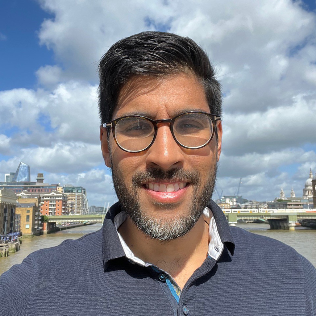 Asian man with glasses and a beard looking straight at the camera with the London skyline behind him