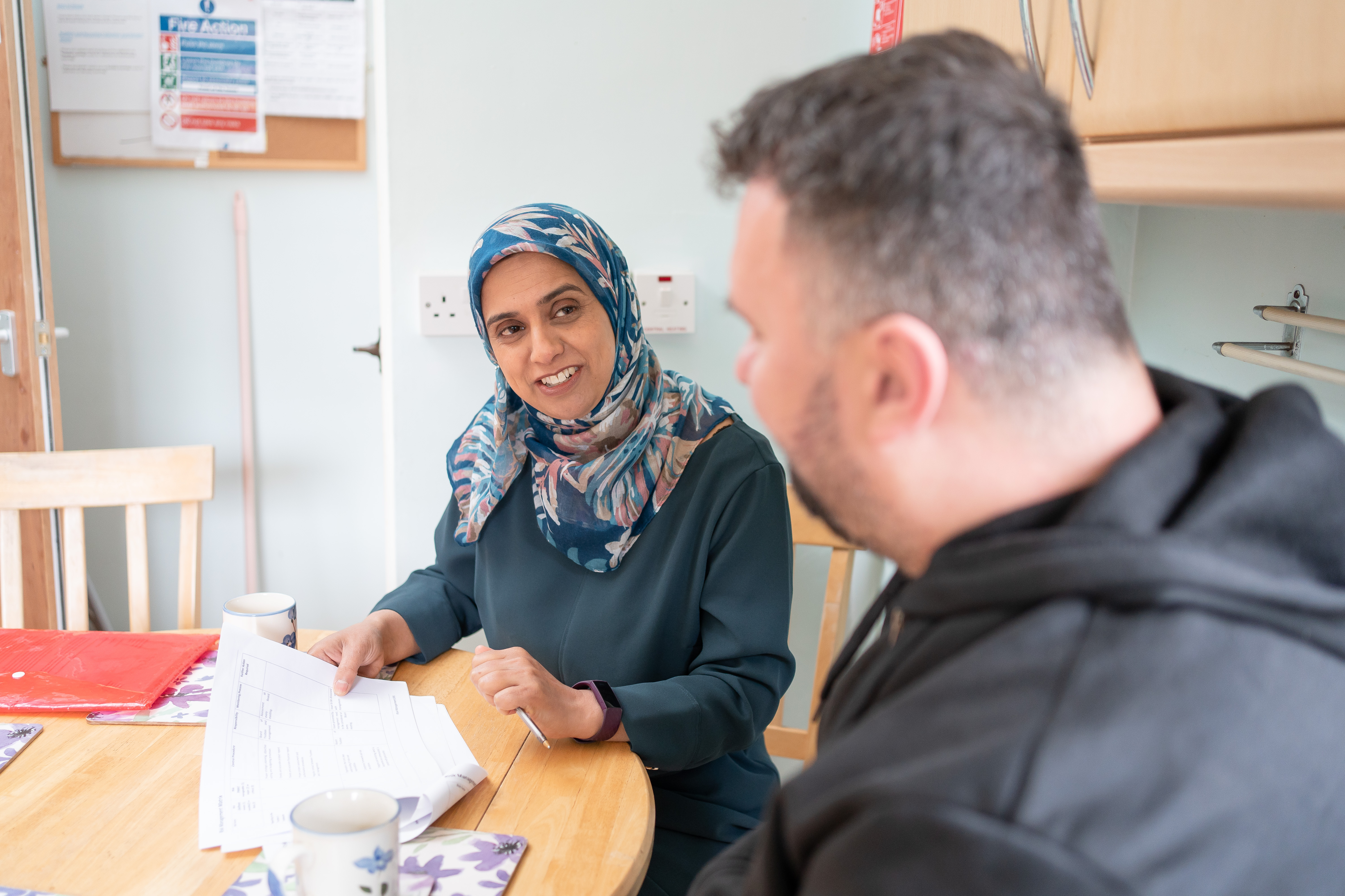 Asian woman in a head scarf talking to a white man with his back to the camera in a kitchen