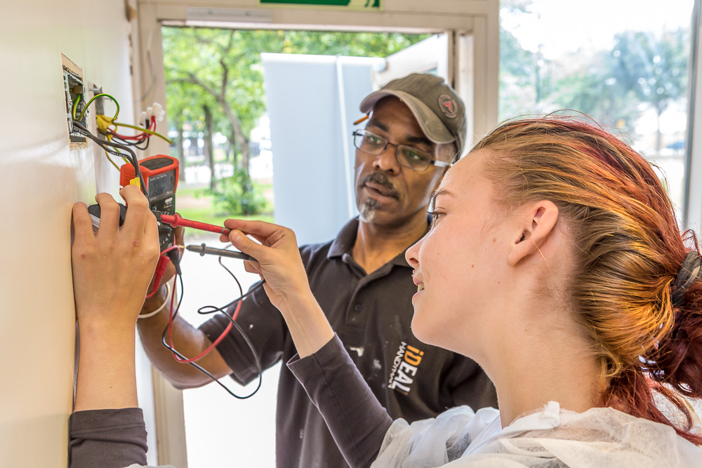 A black man shows a young white girl how to mend a fuse cupboard