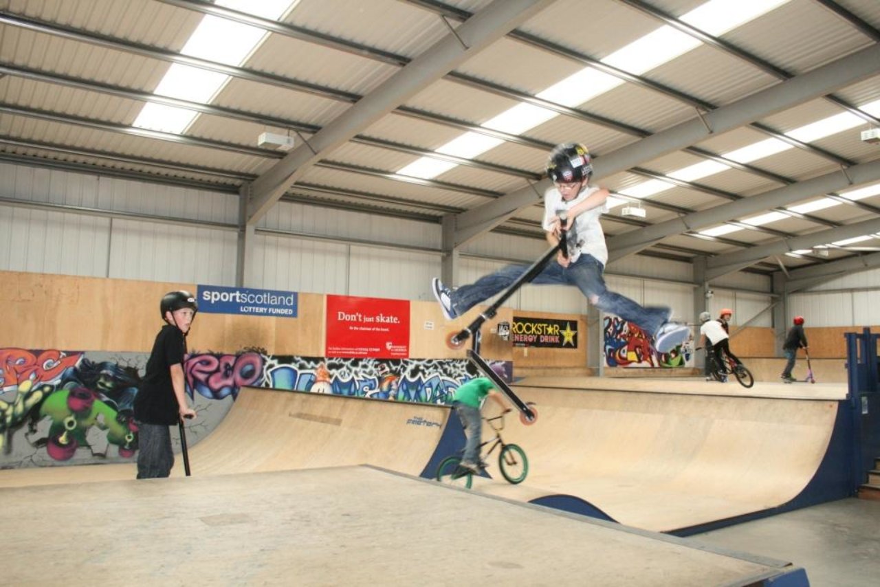 A boy jumping in the air off his scooter with other boys watching him