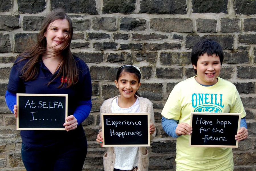 SELFA CEO and two children holding a series of chalkboards that say 'At SELFA experience happiness, have hope for the future.'
