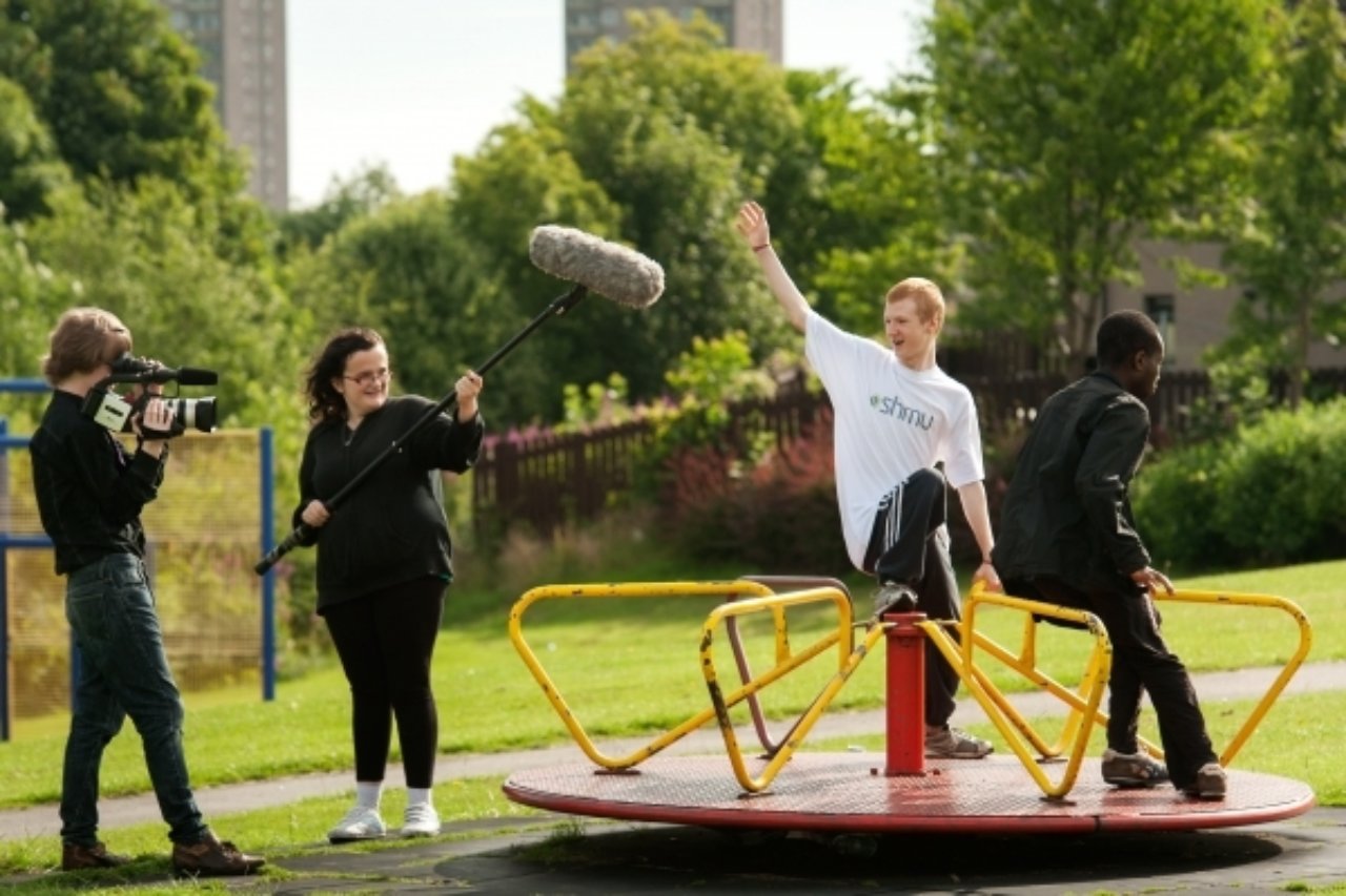 Two boys sitting on a children's roundabout being filmed