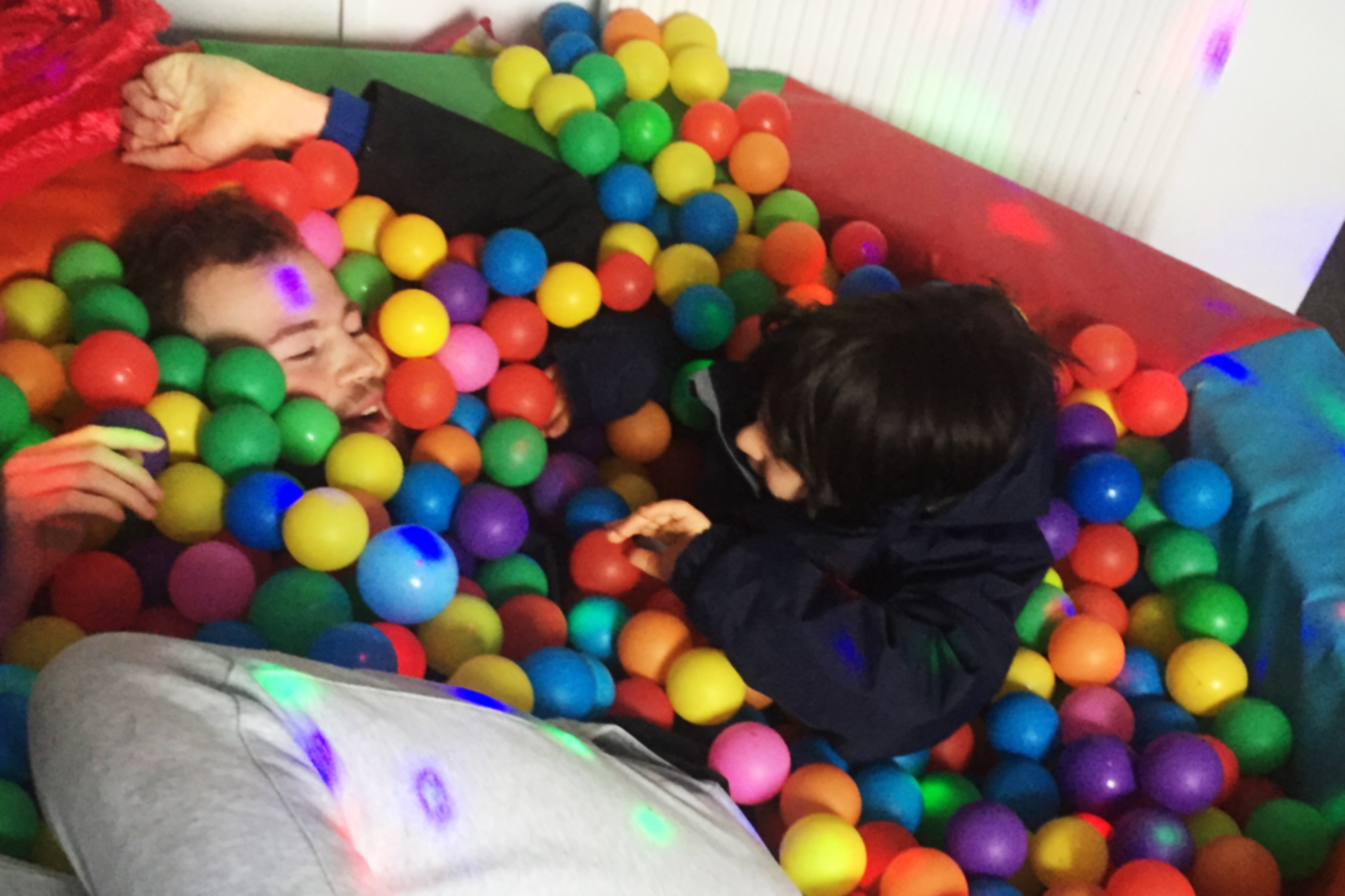 Young white adult laying down in a ball pit with a younger child next to them