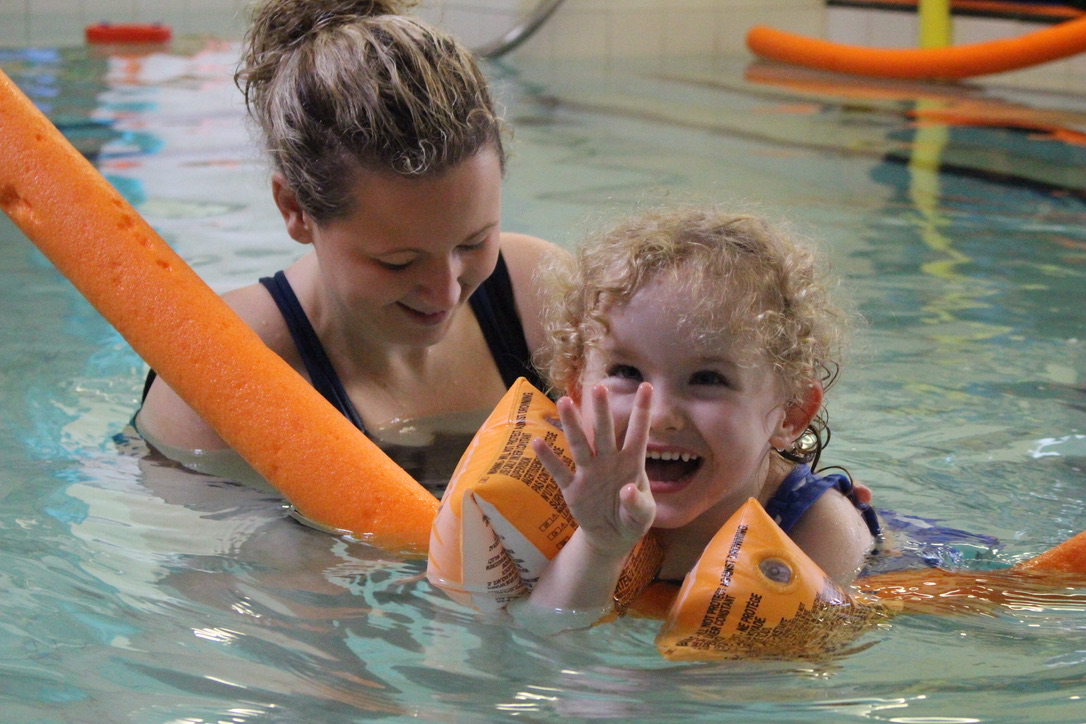 Blonde toddler wearing inflatable orange armbands and a pool noodle being held by a white woman in a swimming pool