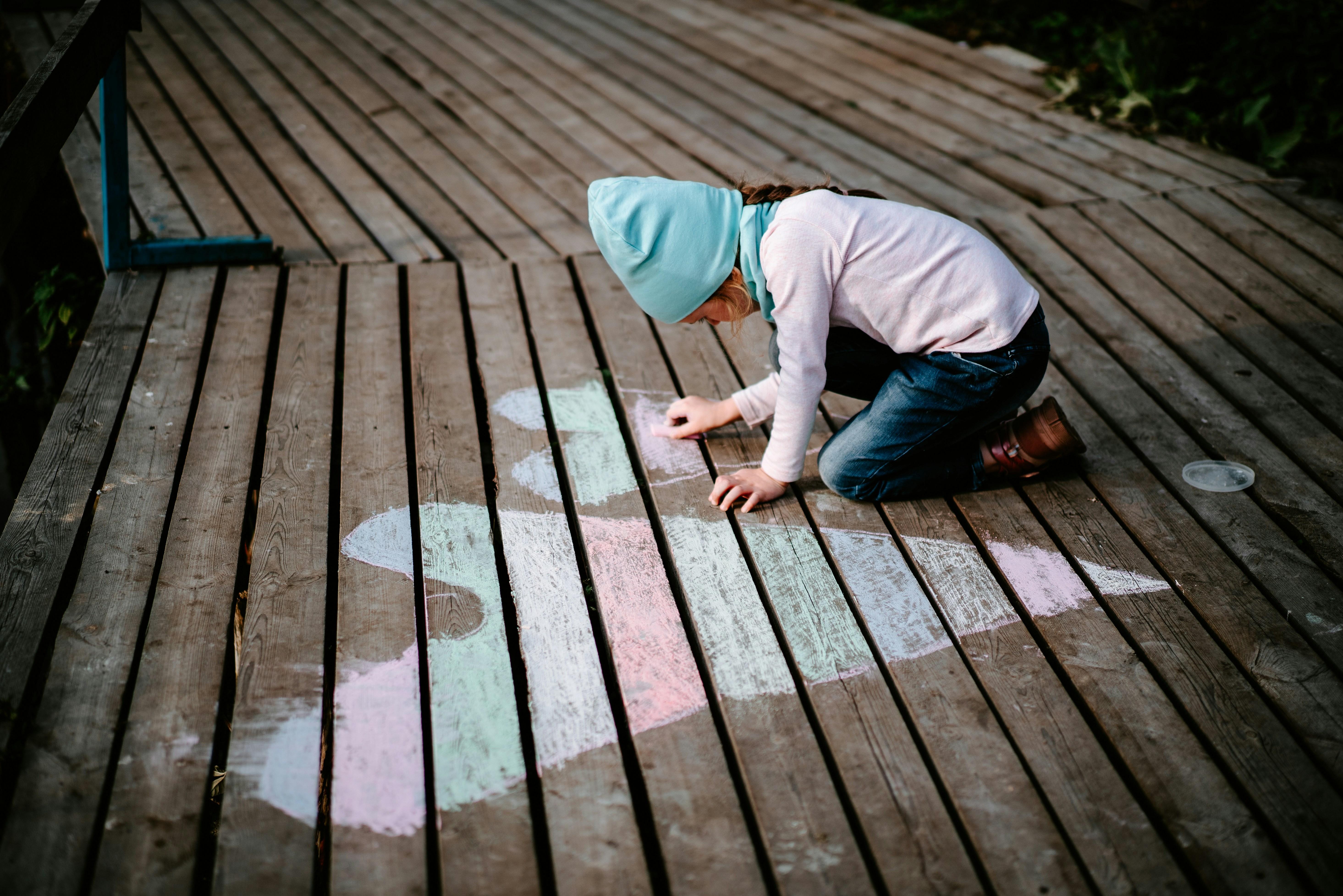 A young child kneels on decking, drawing stripped hearts in pastel colours with chalks, she is wearing a turquoise beany, jeans and a pastel purple long sleeve top